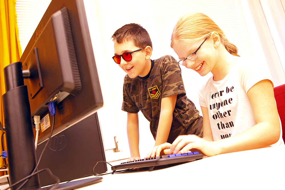 Eine Schülerin sitzt vor einem PC-Bildschirm und schreibt auf der Tastatur. Daneben steht ein Mitschüler und schaut auf den Bildschirm, beide lächeln.