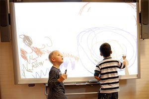 Zwei Grundschüler an einem interaktiven Whiteboard, der eine malt, der andere hält einen Stift in der Hand und schaut nach oben zum Beamer.
