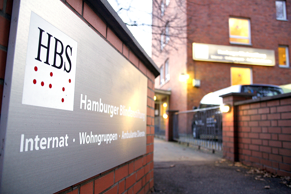 Eingang zum Internat mit einem Schild im Vordergrund auf dem steht: HBS, Hamburger Blindenstiftung, Internat, Wohngruppe