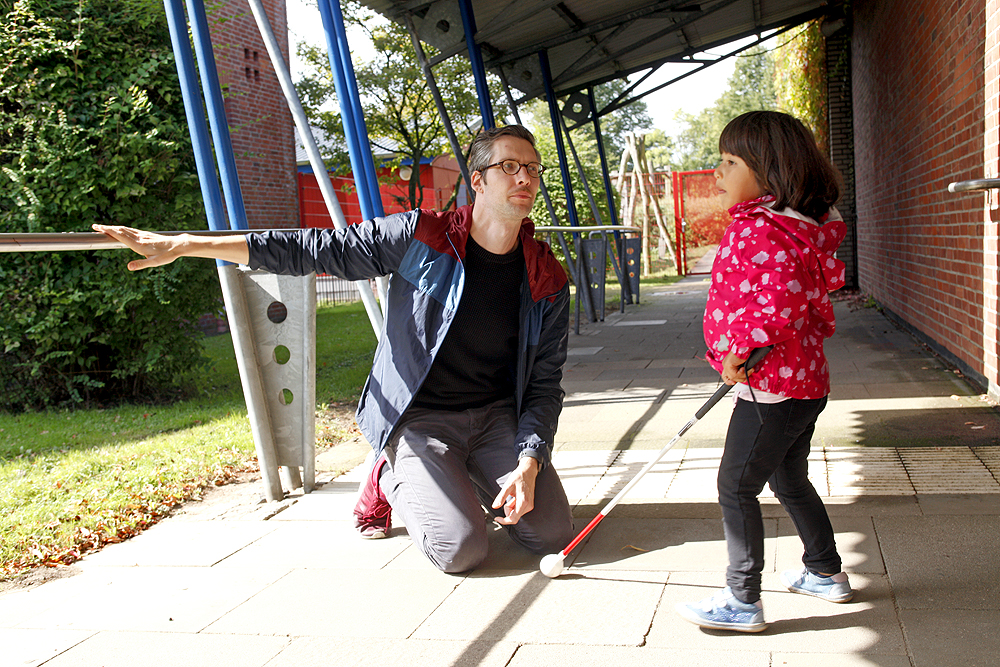 Ein Pädagoge zeigt einem blinden Mädchen das Geländer-Leitsystem im Außenbereich.